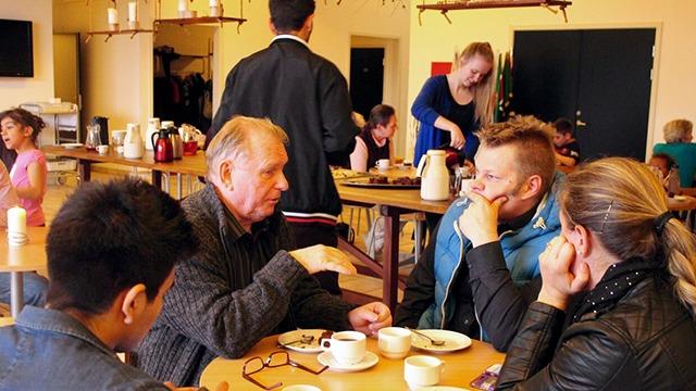 Mennesker sidder ved et bord og snakker over kaffe og te i en hyggelig café. Baggrunden viser flere gæster.