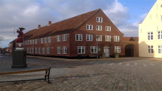 Rød murstensbygning med mange vinduer og en statue på en plads under en blå himmel.