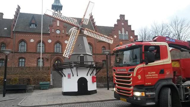 En rød brandbil parkeret foran en vindmølle og en bygning i baggrunden.