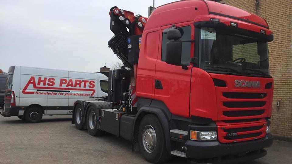 Rød Scania lastbil med kran og AHS Parts varevogn i baggrunden på en gårdsplads.