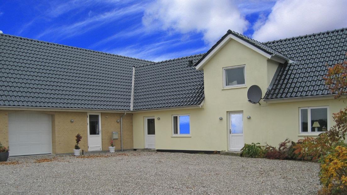 Et moderne hus med grå tag og gule vægge, beliggende på en grusvej under en blå himmel.
