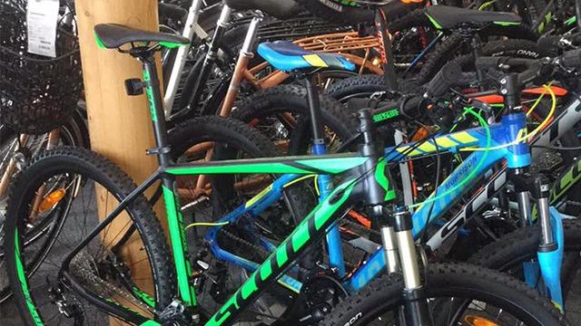 Flere mountainbikes i forskellige farver, herunder grøn og blå, står opstillet i en cykelbutik.