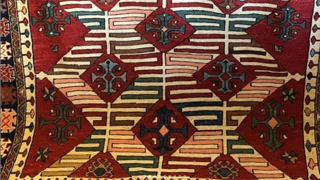 Farverigt tæppe med geometriske mønstre i røde, grønne og gule nuancer. Designet inkluderer diamanter og linjer.