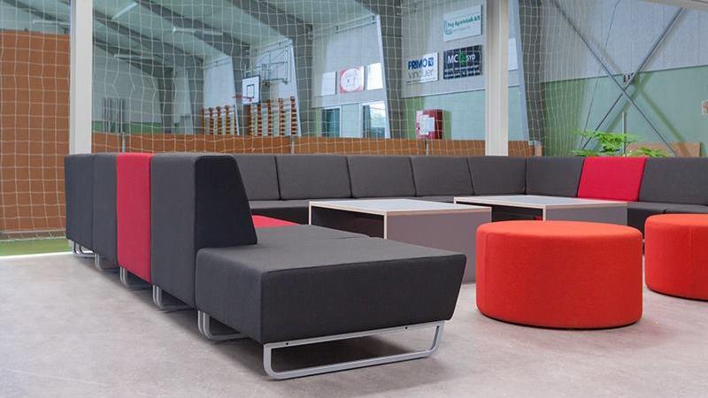 Moderne loungeområde med grå og røde sofaer samt runde borde i en sportsfacilitet.