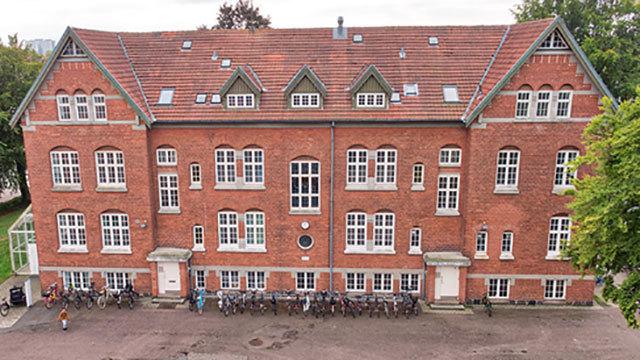 En rød murstensbygning med mange vinduer og cykler parkeret foran. Træer ses i baggrunden.