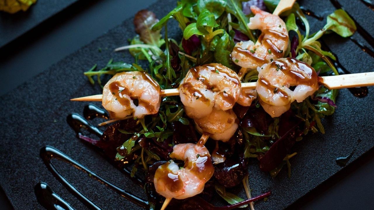 Grillede rejer på spyd serveret med frisk salat og balsamico sauce på en sort tallerken.