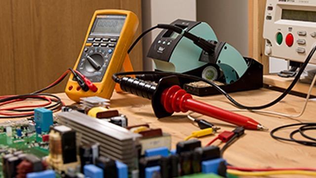 Et arbejdsbord med et multimeter, loddekolbe og elektroniske komponenter til reparation.
