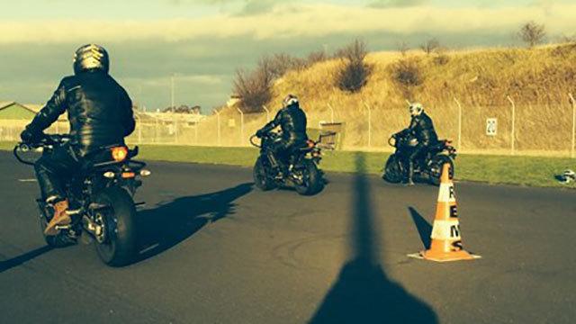 Tre motorcyklister kører på en bane, mens de navigerer omkring en trafikcone i sollys.