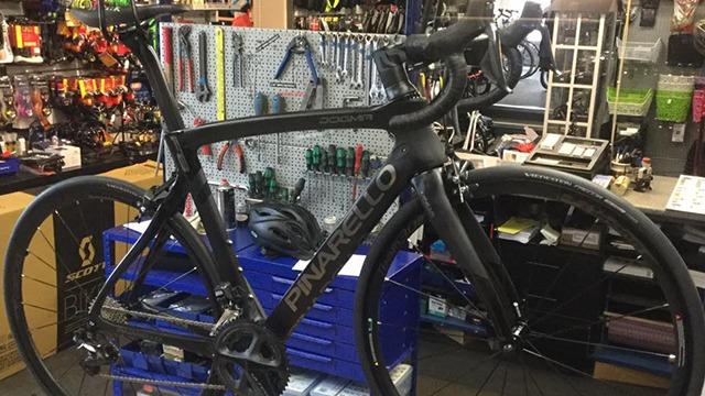 En sort racercykel står i en cykelbutik med værktøj og tilbehør i baggrunden.