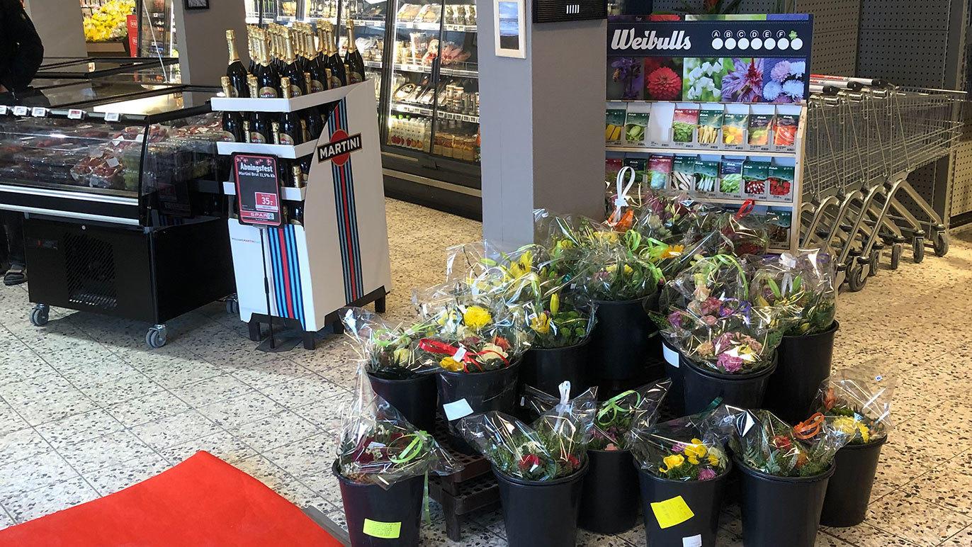 Blomsterarrangementer i sorte potter ved indgangen til en butik, med champagne og frugtjuice i baggrunden.