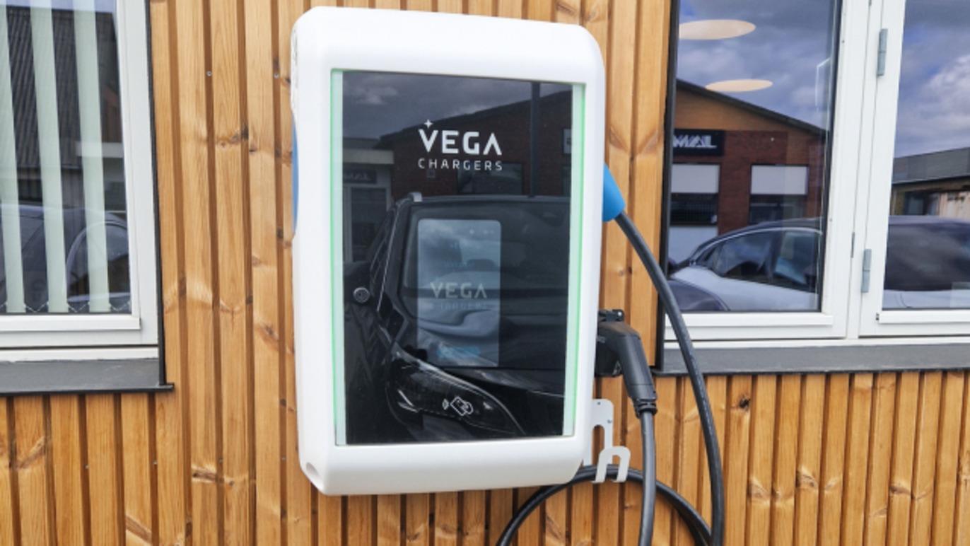 Ladepunkt til elbiler med VEGA-logo, monteret på en trævæg, med ladekabel tilsluttet.