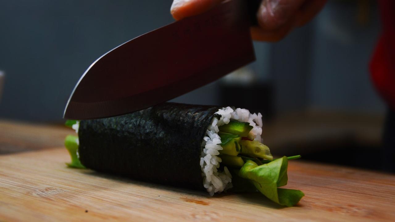 En hånd skærer en sushi-rulle med grøntsager og ris på et skærebræt.