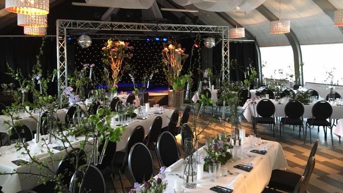 Elegant festlokale med dækkede borde, blomsterdekorationer og en scene med lys og pynt.