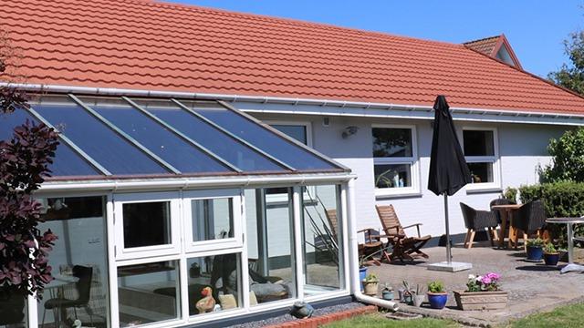Et hus med en glasvæg, terrasse og havemøbler under en parasol på en solrig dag.