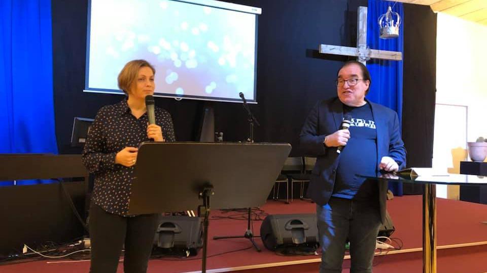 To personer taler ved en prædiken i en kirke med en kors og skærm i baggrunden.