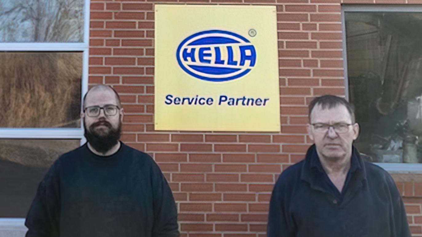 To personer står foran en HELLA servicepartner-skylt på en murstensvæg.