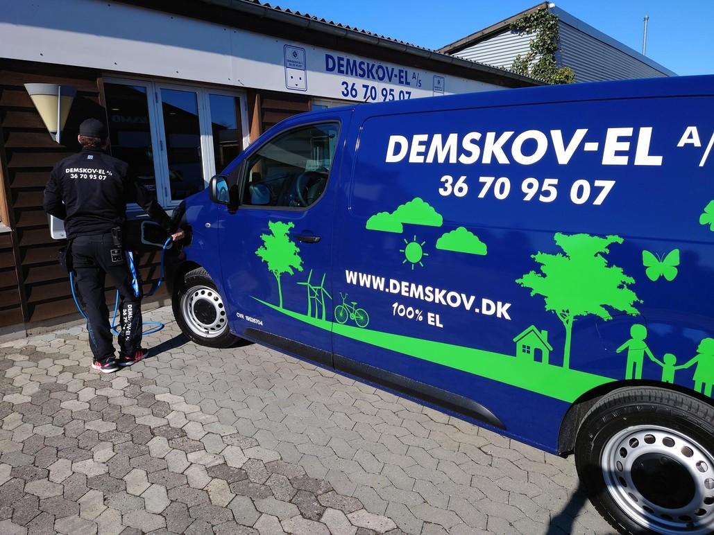 En mand står ved en blå varevogn med teksten "DEMSKOV-EL" og kontaktinformationer, foran en bygning.