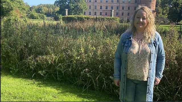 Kvinde står i en have med høje græsser og en bygning i baggrunden på en solrig dag.