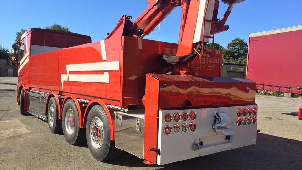 Rød lastbil med kran og trailer, set fra siden, parkeret på en gårdsplads.