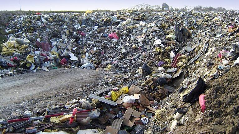 Affaldsdeponi med bunke af skrald, herunder plastik, pap og tekstiler, ved en grusvej.