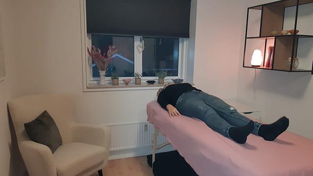 En person ligger på en massagebriks i et lyst rum med planter og en stol i baggrunden.