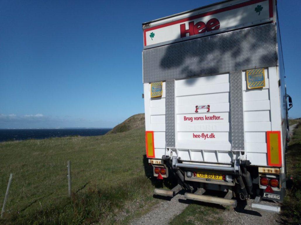 Lastbil med logo ved kysten, der kører på en grusvej med udsigt til havet.