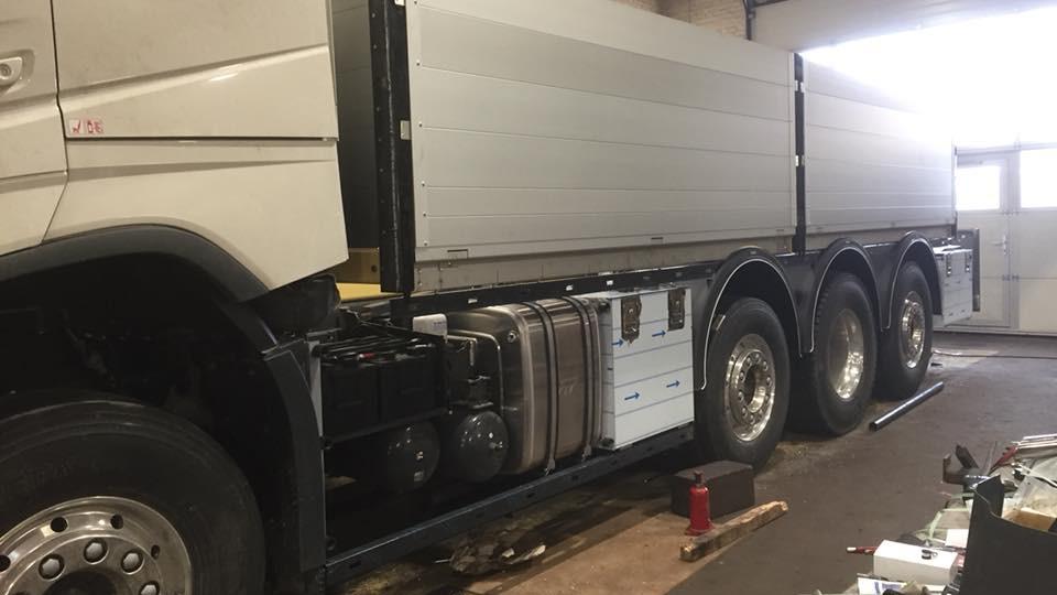 En stor lastbil med tre aksler står i en garage, med synlige brændstoftanke og en hvid kasse.