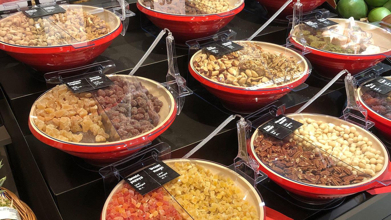 Skåle med forskellige nødder og tørret frugt i en butik, præsenteret i røde skåle med labels.