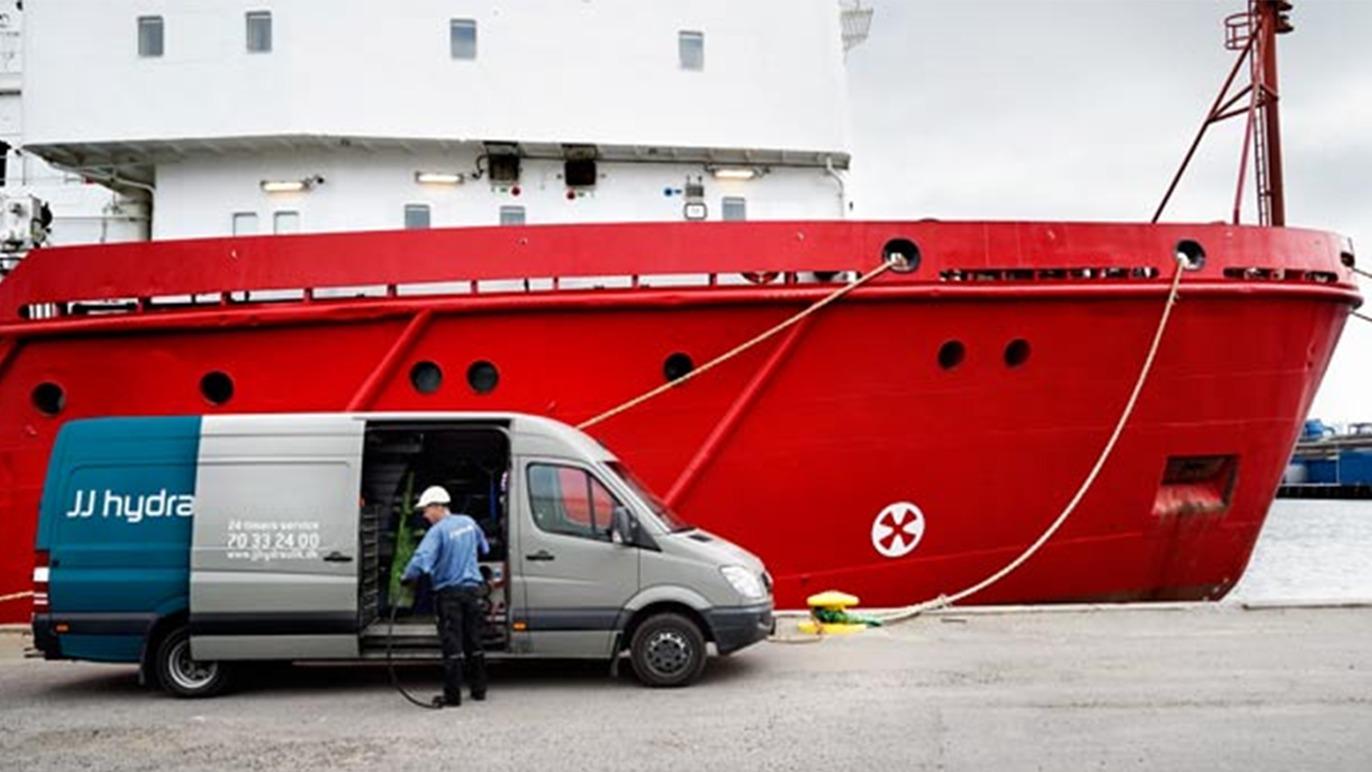 En grå varevogn står ved siden af et stort rødt skib i havnen, mens en mand arbejder ved åbne døre.