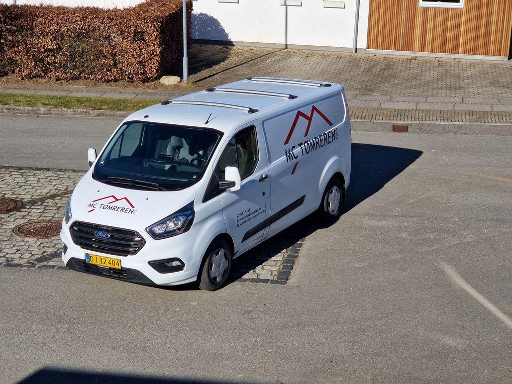 En hvid varevogn med logo parkeret på en grusvej, omgivet af buske og bygninger.