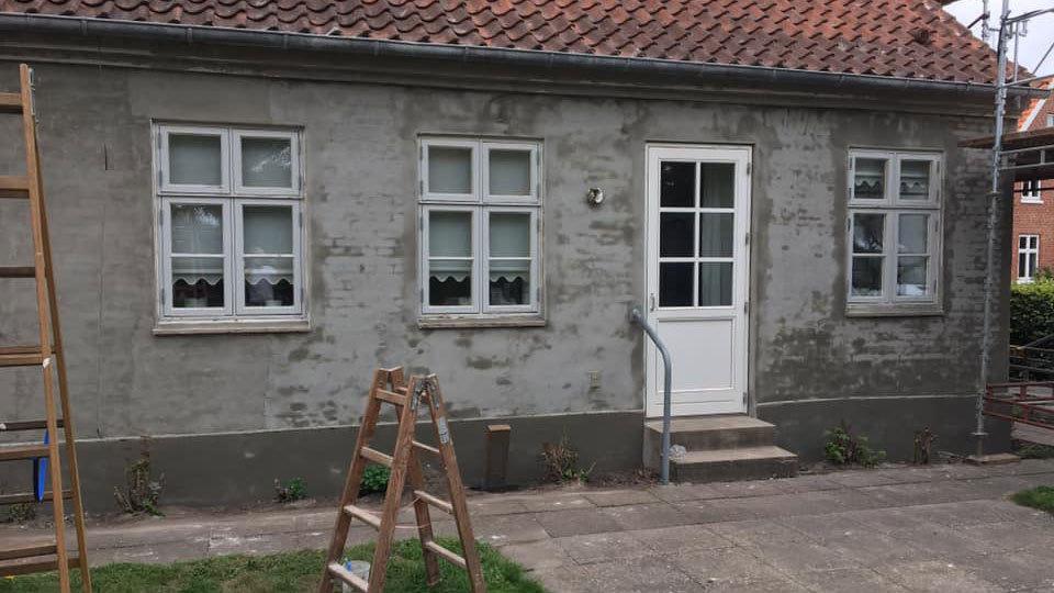 En grå væg af et hus med hvide vinduer og en hvid dør, samt en stige i forgrunden.