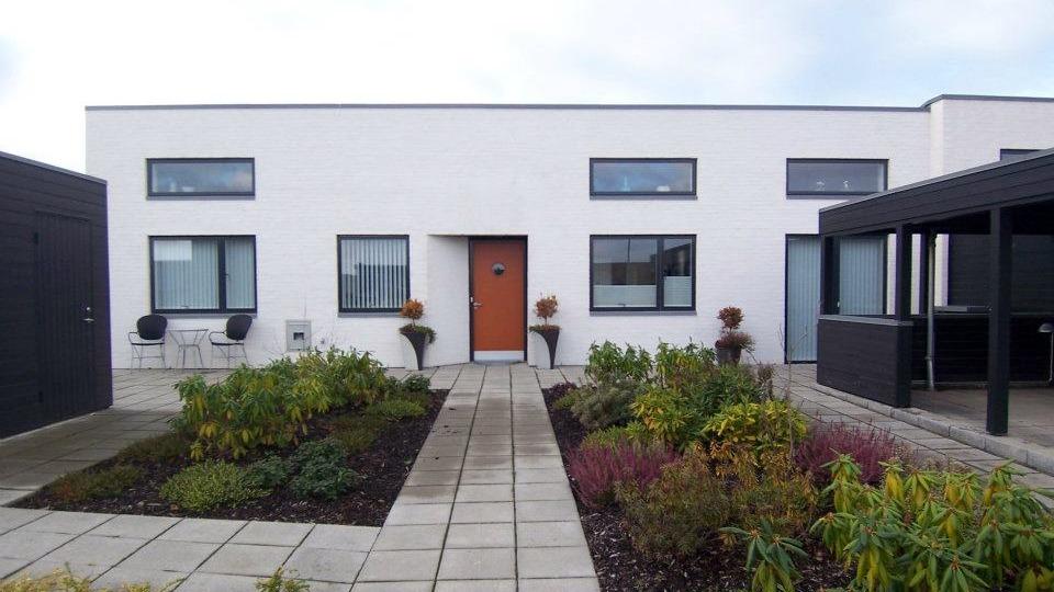 Moderne hvidt hus med orange dør og haveanlæg foran, omgivet af fliser og planter.