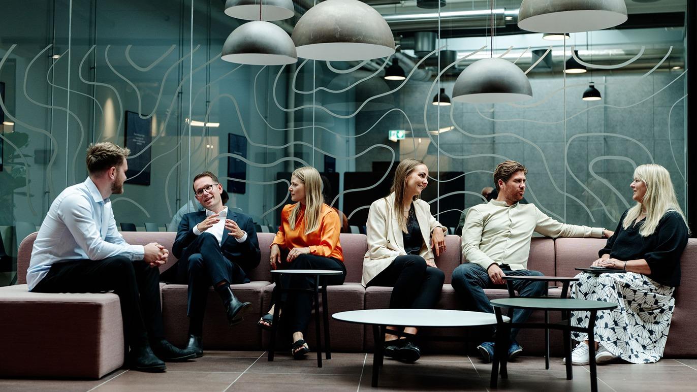 Gruppe af seks personer, der sidder i en moderne lounge med sofaer og lamper, mens de taler sammen.