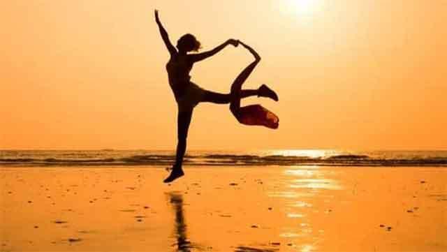 En person danser på stranden ved solnedgang, med havet i baggrunden og en gylden himmel.