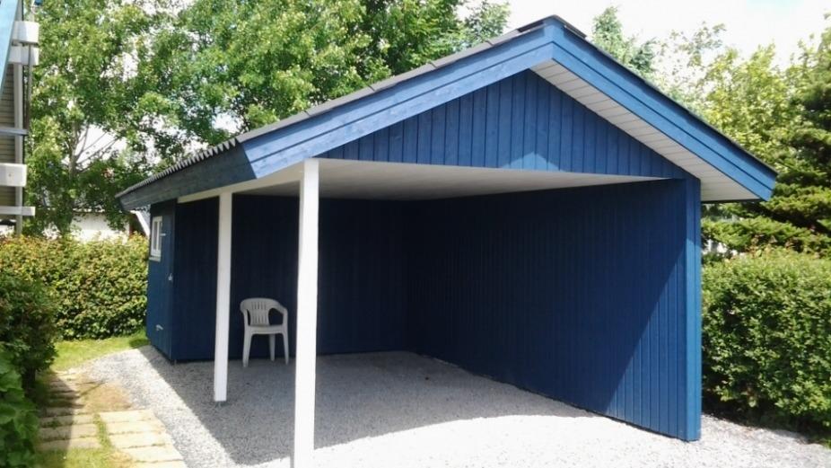 Blå carport med grå grusbelægning og en hvid stol under taget, omgivet af grønne buske.