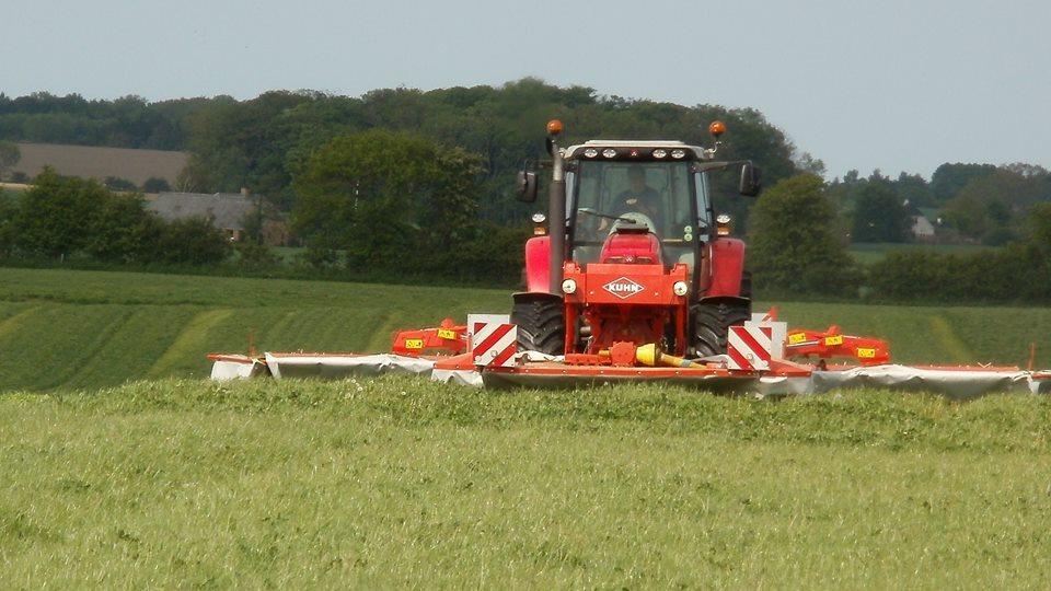 Rød traktor, der klipper græs på en mark med grønne marker i baggrunden.
