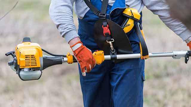 En person holder en græstrimmer med orange handsker og blå arbejdstøj i en udendørs have.