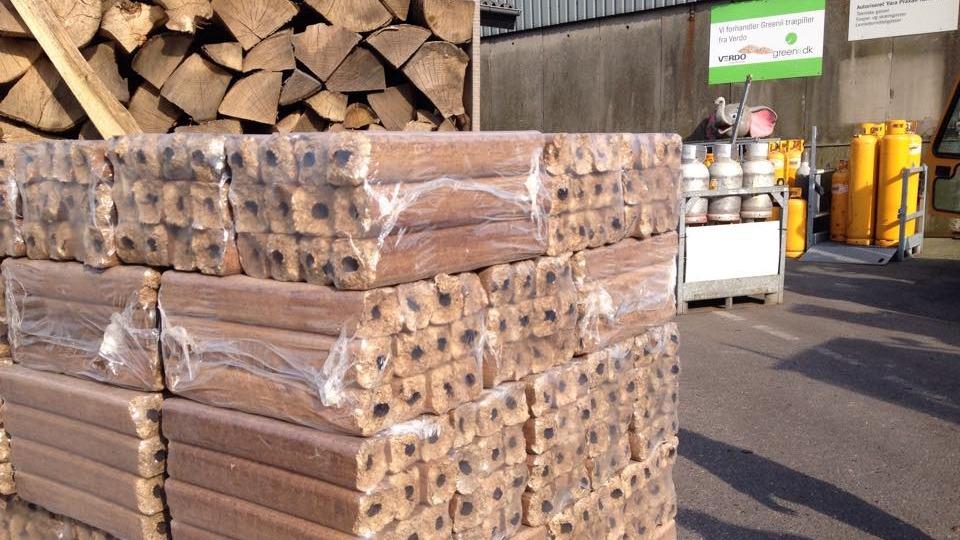 Stak med brændstoftræ pakket i plastfolie ved en bygning med gasflasker i baggrunden.