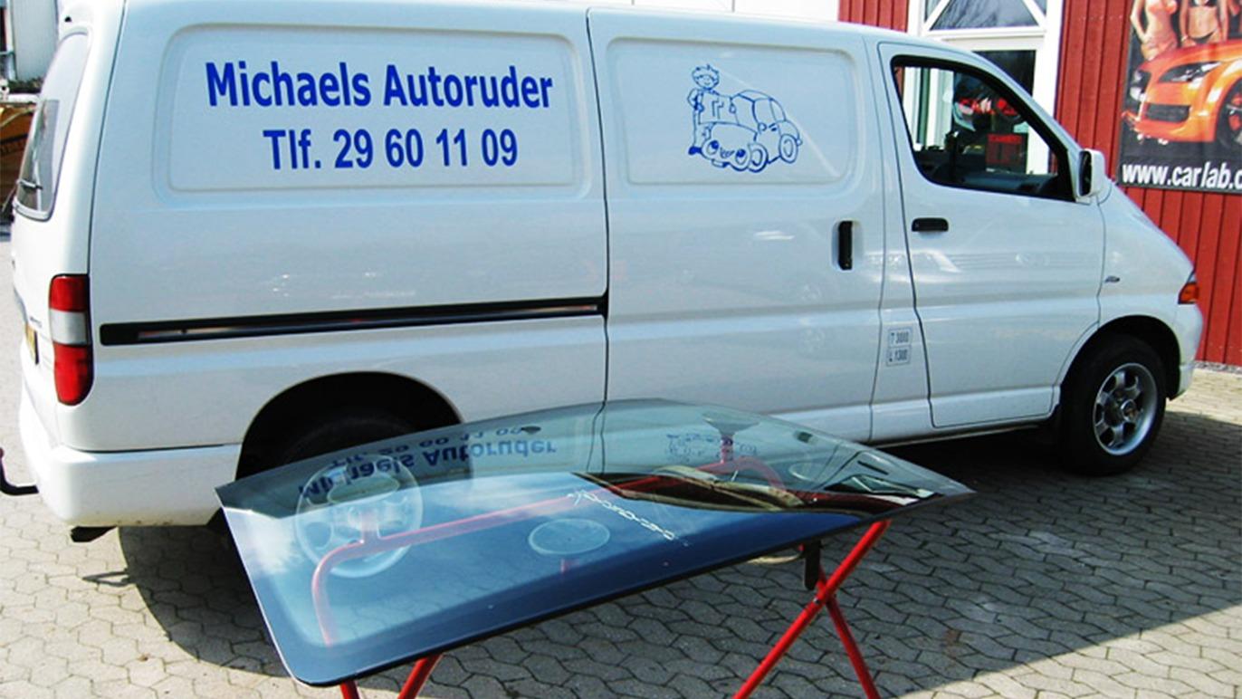 En varevogn med teksten "Michaels Autoruder" og en bilrude på et stativ i forgrunden.