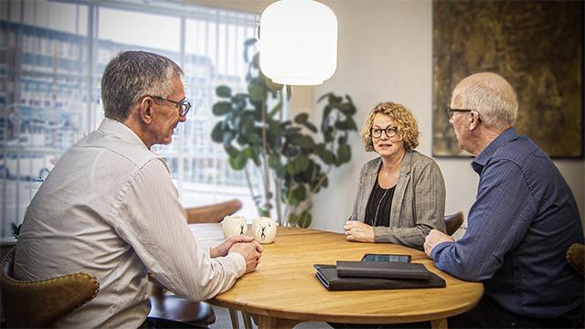 Tre personer sidder ved et rundt bord i et lyst rum, mens de diskuterer med kaffekopper i hånden.