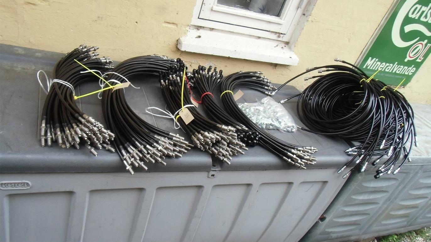 Bunke af sorte kabler samlet på en grå kasse ved en væg, med nogle metaldele i en plastikpose.