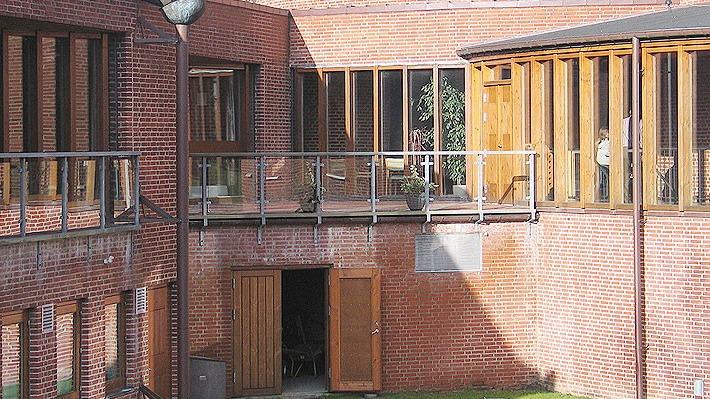 En gård med murstensvægge og store vinduer, der giver udsigt til en indendørs terrasse og grøn græsplæne.