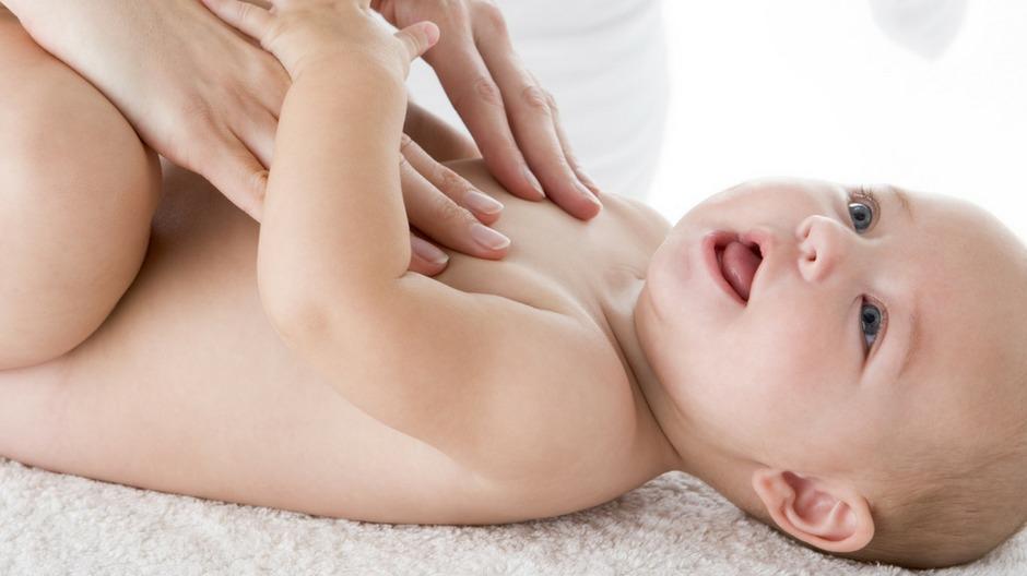 En baby ligger på en blød overflade, mens en voksen masserer dens mave med blide hænder.
