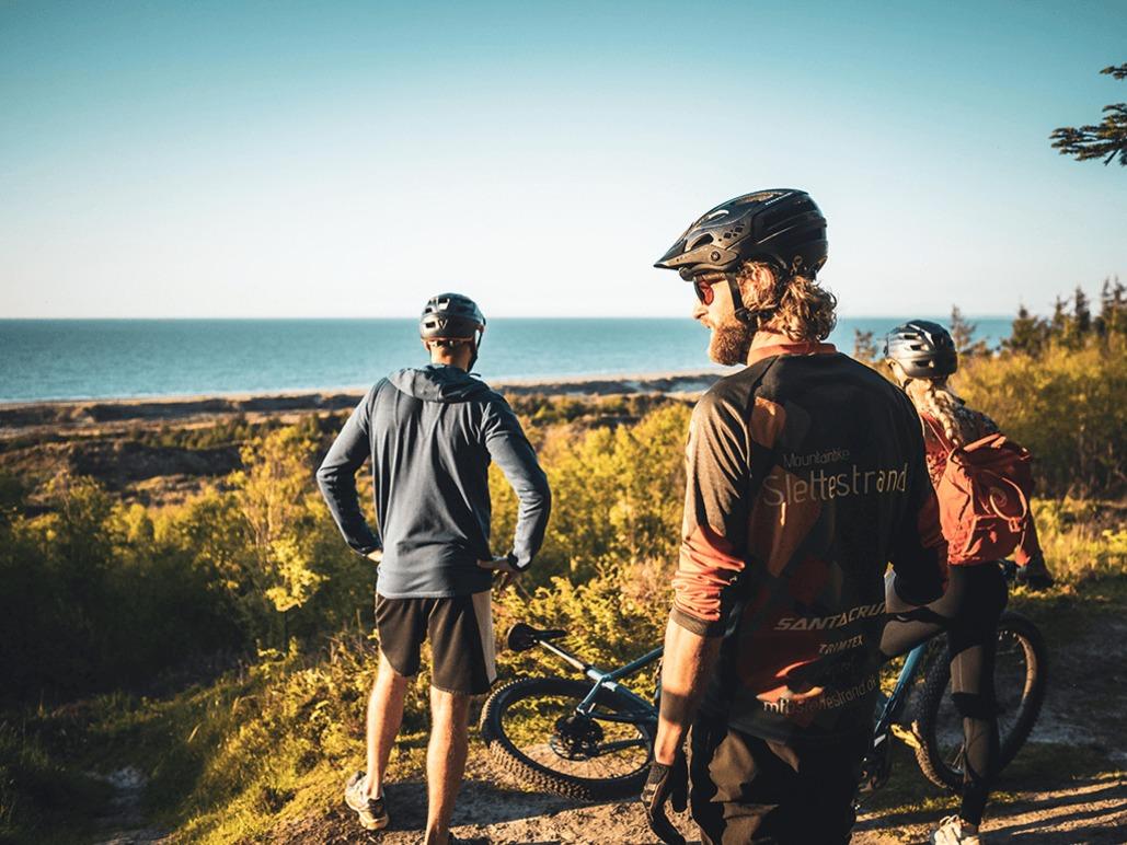 Tre mountainbikere står på en sti med udsigt over havet og nyder den smukke natur.