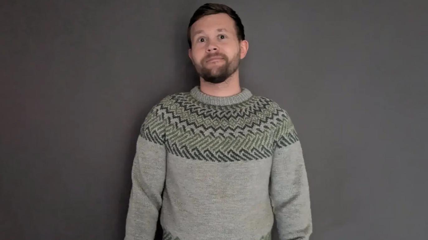 Mand iført en grå sweater med mønster, står foran en grå væg og ser direkte ind i kameraet.