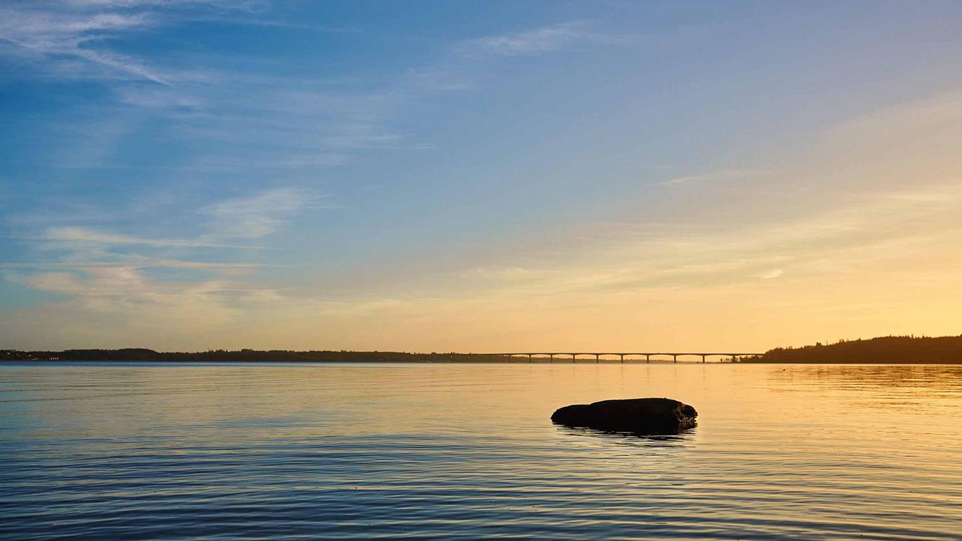 En stille sø med en stor sten i forgrunden og en bro i baggrunden under en farverig solnedgang.