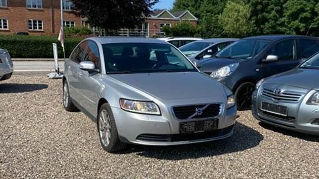 En sølvfarvet Volvo sedan parkeret blandt andre biler på en grusbelagt parkeringsplads.