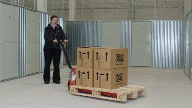 Kvinde bruger en palleløfter til at flytte kasser i et lagerområde med metalvægge.
