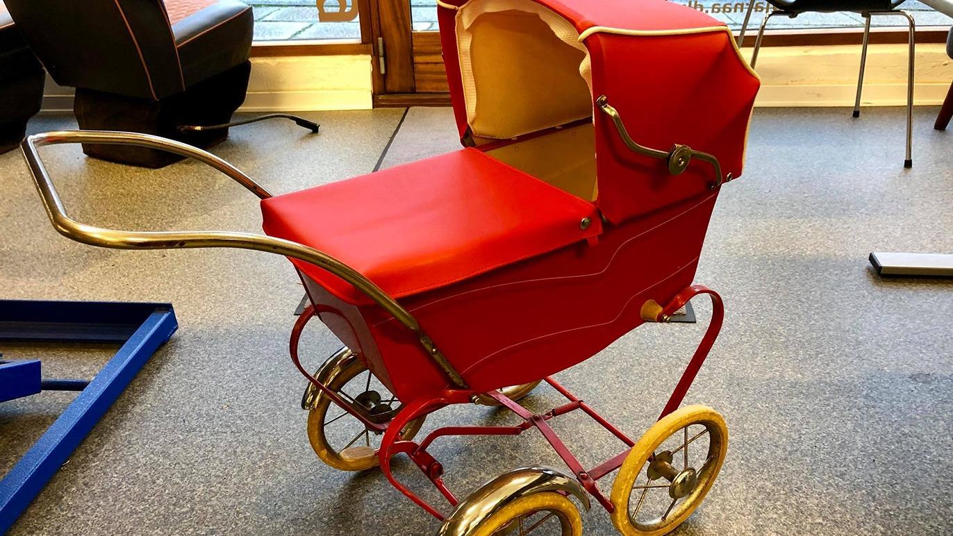 En rød dukkevogn med metalramme og gule hjul står på et gulv i et rum.