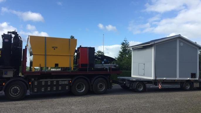 Lastbil med en gul container og et lille hus på en trailer, parkeret på en grusvej under en blå himmel.